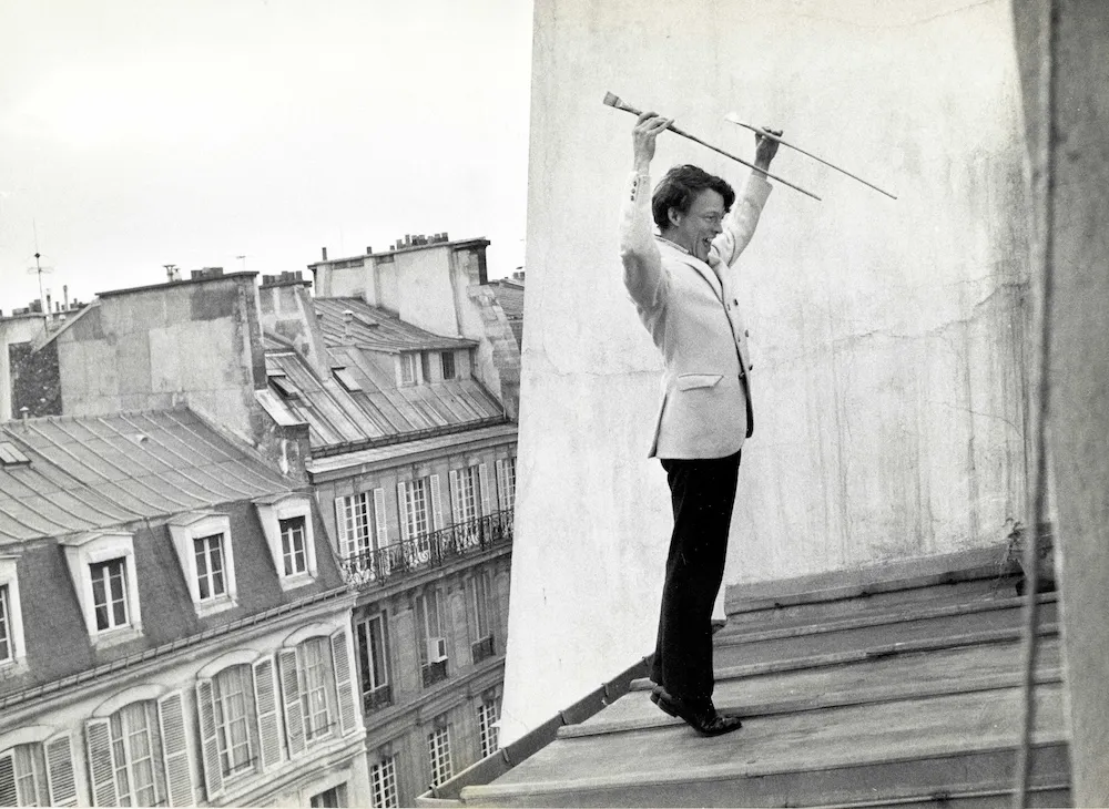 A man in a white jacket and dark pants stands on a roof with two spears in his hand like a Picador.