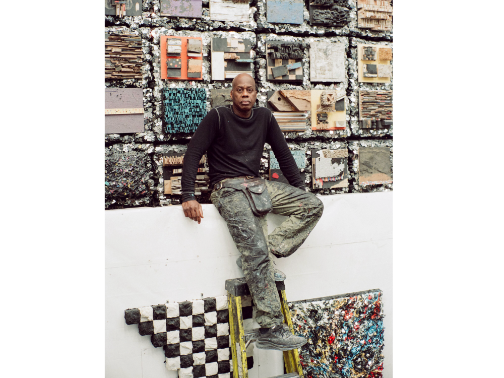 A man in a dark shit and paint covered chinos stands on a ladder. he is surrounded by artworks.