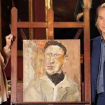 A man in a blue suit and a women in a leather jacket flank a smallish square painting of a man in a black scarf.