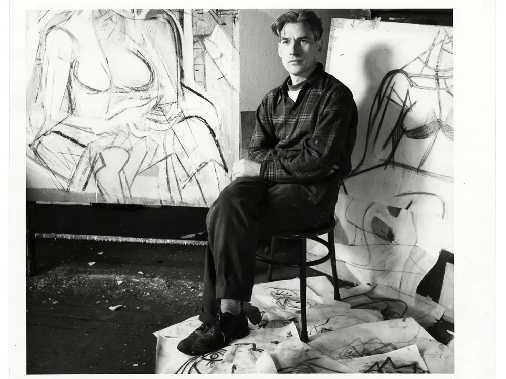 black and white photo of a young man sitting on a chair, with charcoal drawings scattered on the floor and two large drawings propped up against the wall behind him.
