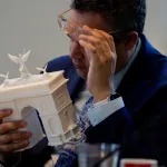 A man in a blue suit jacket lifts his glasses to inspect a model of a
