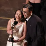 A man in a white dress and a man in a tuxedo holding a golden statuette and smiling before a microphone.