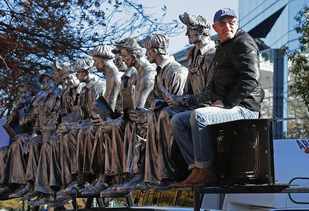 Sergio Furnari sits beside his large-scale sculpture inspired by the iconic 1932 photograph Lunch Atop a Skyscraper, depicting a row of ironworkers seated high above Manhattan, during a public installation in New York City.