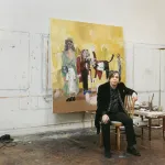 Portrait of George Condo in his studio with a painting behind him.