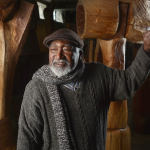 Portrait of Thaddeus Mosley wearing a scarf with one hand on a carved wooden sculpture.