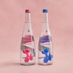 two clear glass bottles in front of a light pink backdrop. The one on the left has an image of a pink balloon dog on the label, and the one on the right has a blue balloon dog.