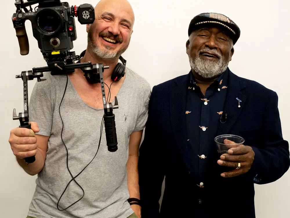 Two men standing side by side and smiling. On the left is a bald white man wearing a gray t-shirt and green pants, with a large camera resting on his shoulder and headphones around his neck. On the right is an older black man holding a plastic cup, wearing a navy jacket and a hat.