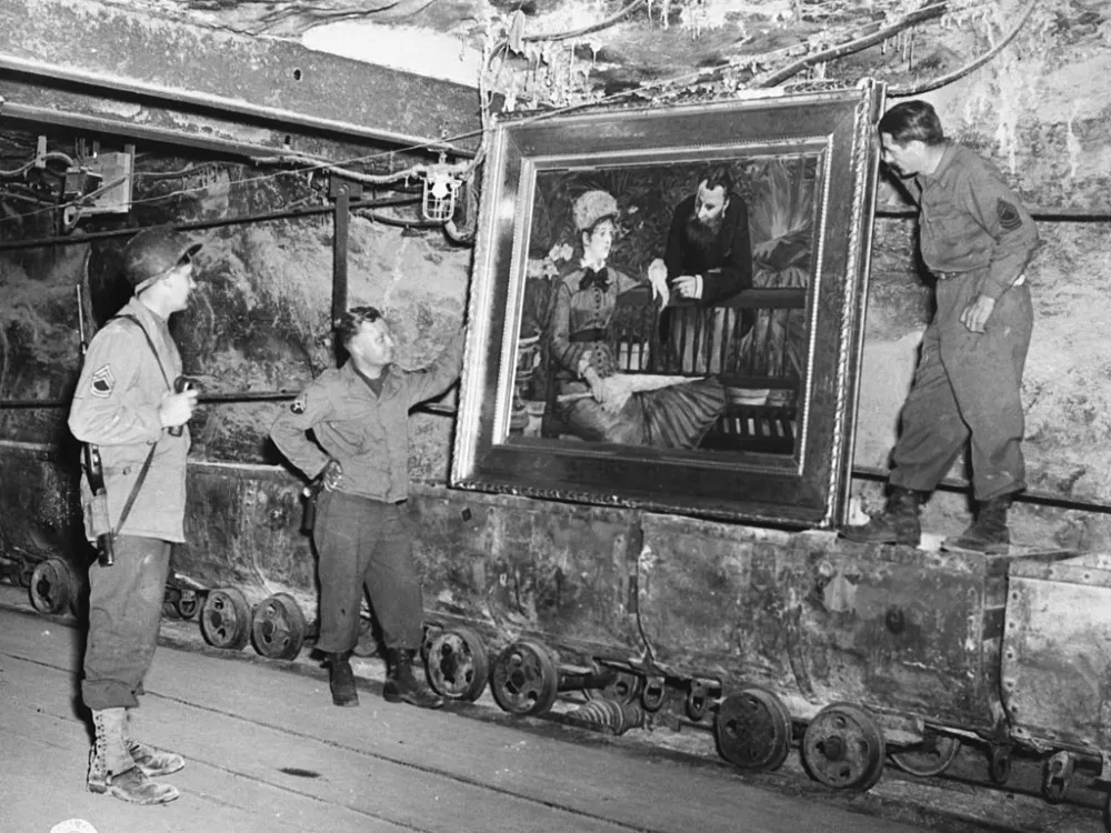 black-and-white photo of three men in army uniforms looking at a framed painting on a rough wall. They