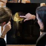 Two women pointing at a canvas painted with an image of a man in golden robes illuminated by a spotlight.