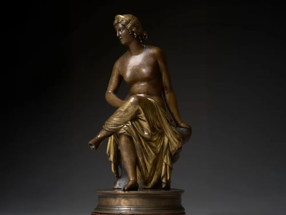 A photo shows a small bronze sculpture of a seated woman, breasts bared, with robes wrapped around her lower half.