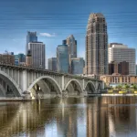 Large buildings reach toward a blue sky in city of Minneapolis behind a peaceful view of St. Anthony Falls.