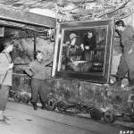 black-and-white photo of three men in army uniforms looking at a framed painting on a rough wall. They