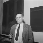 black-and-white photo of a man from the waist up, looking at the camera. He is balding and wearing glasses, a tie, striped collared shirt and suit jacket, holding a cigarette. There are two abstract paintings on the wall behind him.