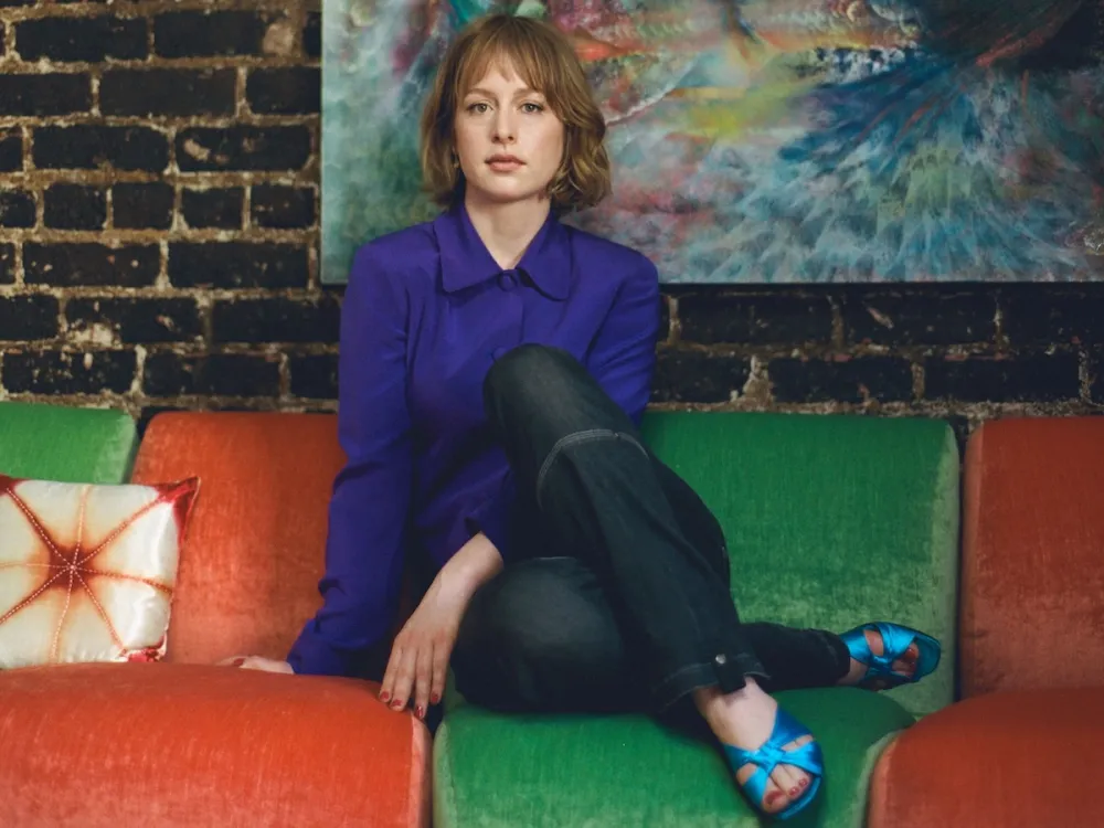 A woman sitting cross-legged on a red and green couch in front of an abstract painting.