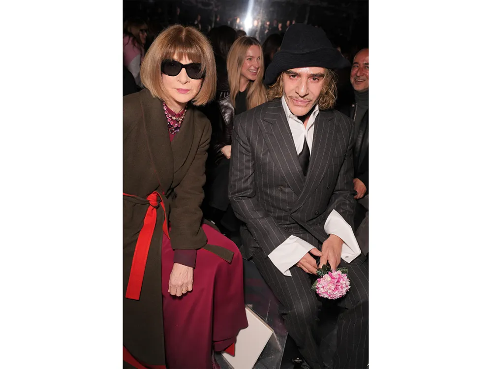 A woman with a severe bob and dark sunglasses wearing a brown coat tied with a red sash at the waist next to a man in a black fedora and pin-stripe suit with white shirt cuffs holding a pink flower